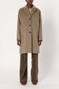 A person wearing a beige manteau mi-long with three buttons, patterned brown pants, and brown shoes stands against a plain white background.