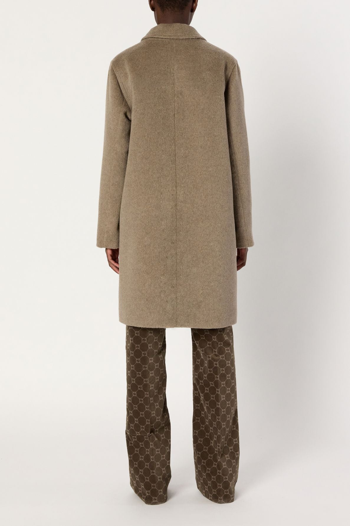 A person stands facing away, wearing a manteau laine mi-long beige over brown pants with a light geometric pattern, against a plain white background.