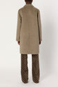 A person stands facing away, wearing a manteau laine mi-long beige over brown pants with a light geometric pattern, against a plain white background.