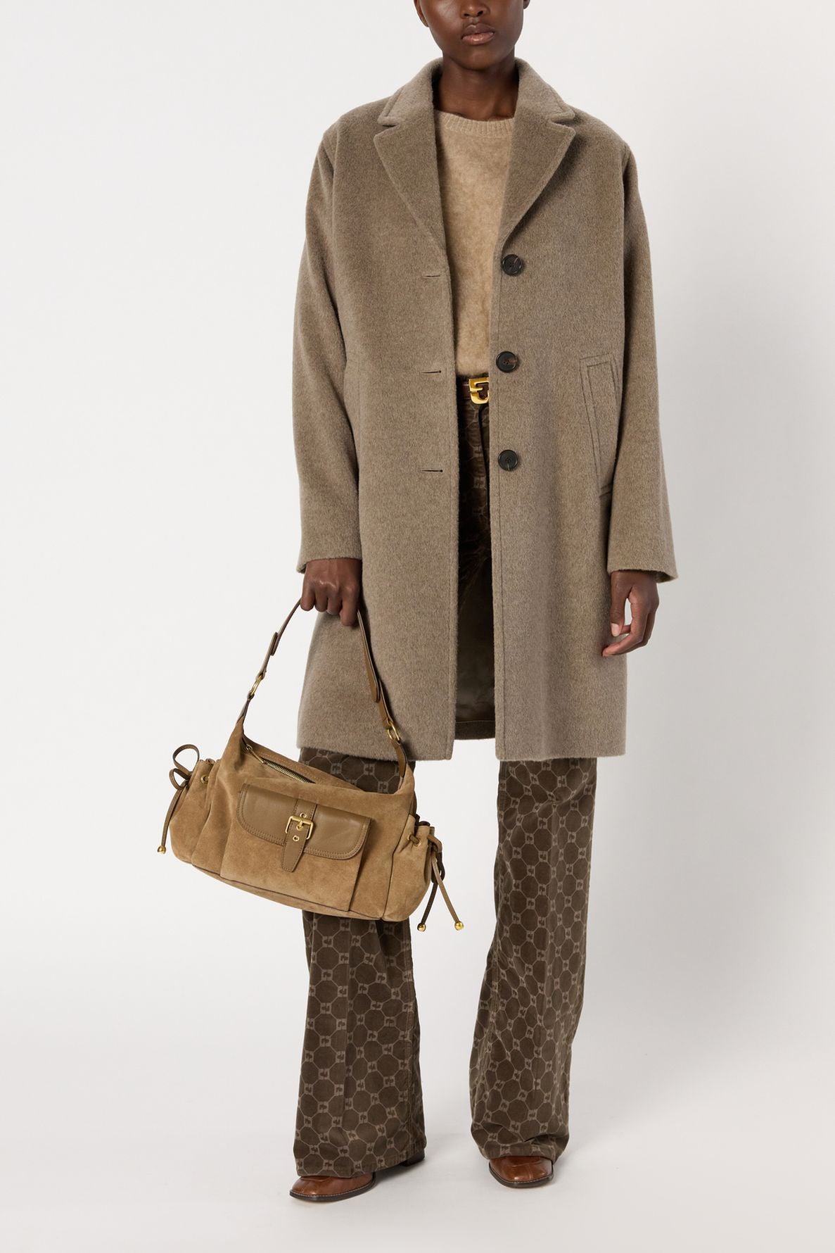 A person wearing a beige manteau laine over a sweater and patterned brown pants holds a tan handbag. They are standing against a plain white background.