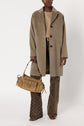 A person wearing a beige manteau laine over a sweater and patterned brown pants holds a tan handbag. They are standing against a plain white background.