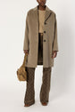 A person wearing a manteau hiver in a tan shade over a beige sweater and patterned brown pants, holding a tan shoulder bag.