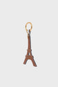 A brown cuir de vachette keychain shaped like the Eiffel Tower, with white stitching details and a gold keyring, embodies élégance parisienne against a plain light background.