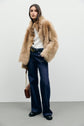 A woman wearing a tan faux fur jacket, white top, and a chic jean flare Anna stands against a light wall, holding a brown handbag with a chain strap. Her black shoes and high-waisted jeans complete the effortlessly stylish look.