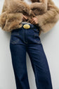 Person wearing a tan faux fur coat and high-waisted dark blue jeans with a wide belt featuring a large oval gold buckle, standing against a plain background. The chic silhouette is enhanced by the jean taille haute for an effortlessly stylish look.