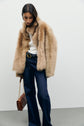 A person wearing a tan faux fur coat, white top, and dark pantalon chic wide-leg jeans stands against a plain light background, holding a brown handbag with a gold chain strap.