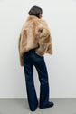 A person with shoulder-length dark hair wears a tan faux fur jacket, dark blue jean taille haute, and black shoes, standing against a plain white background and facing away from the camera.