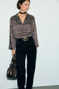 Woman wearing a patterned long-sleeve blouse, black pantalon taille haute with a belt, and a black choker, holding a metallic handbag, standing against a plain light-colored wall.