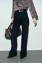 Person wearing a patterned long-sleeve blouse, a pantalon taille haute noir, and black pointed shoes; holding a black handbag with gold details. Only the body from shoulders down is visible.
