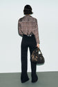 A person stands facing a wall, wearing a patterned long-sleeve shirt, black jean noir taille haute, and holding a large brown handbag.