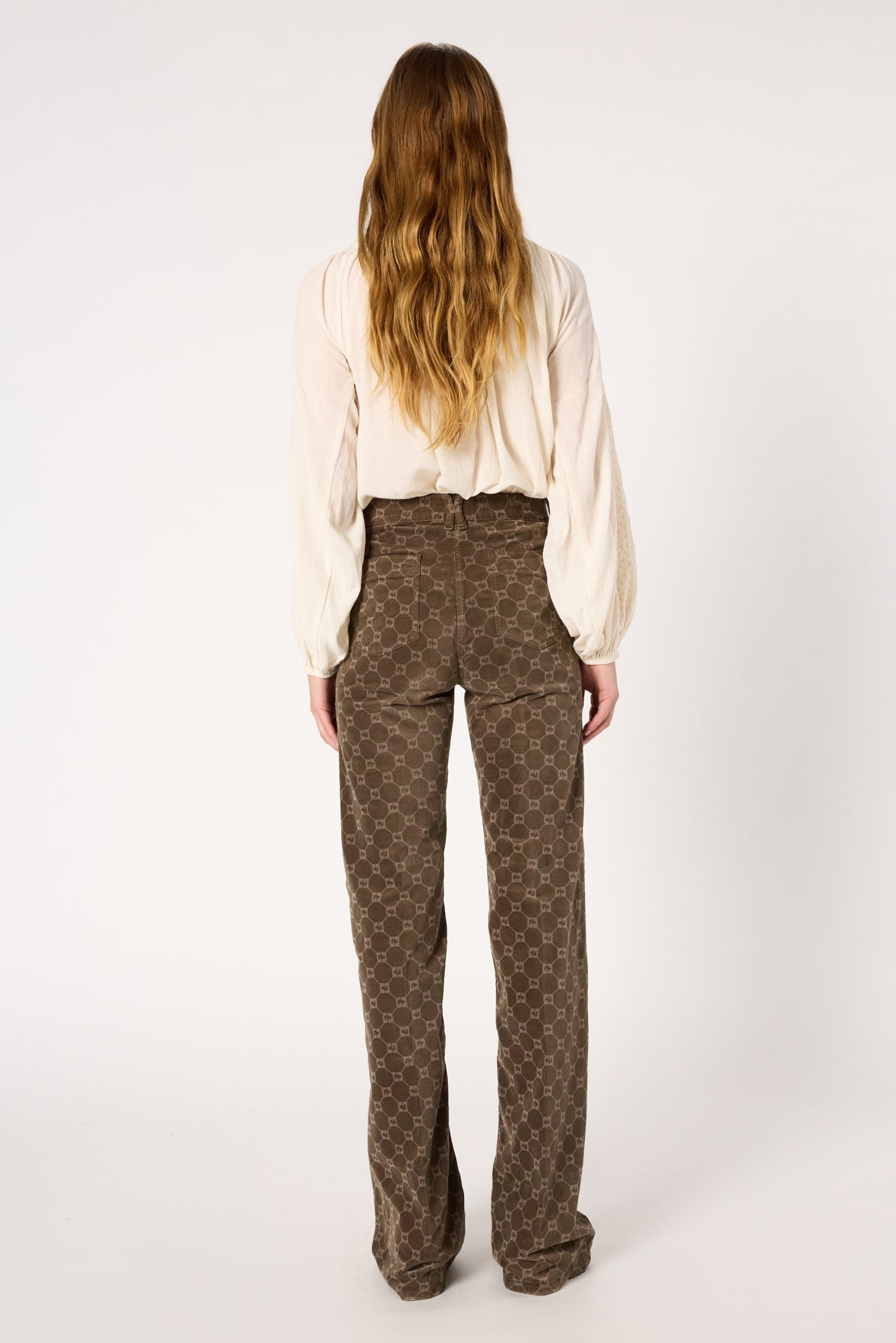 A person with long wavy hair stands facing away, wearing a cream blouse with billowy sleeves and high-waisted brown pants with a geometric pattern. The background is plain white.