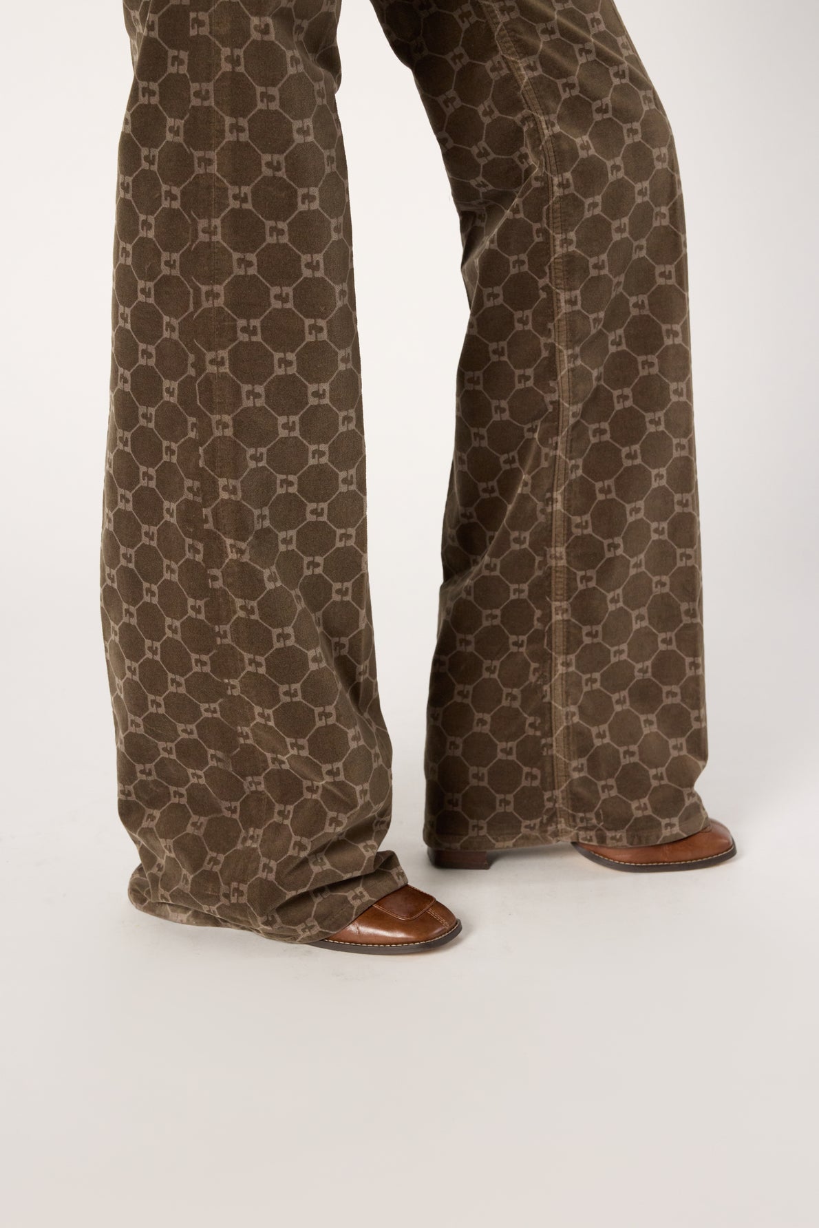 Person wearing wide-legged, patterned brown pants that reach the floor, partially covering brown pointed-toe shoes, standing against a plain white background.