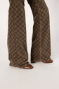 Person wearing wide-legged, patterned brown pants that reach the floor, partially covering brown pointed-toe shoes, standing against a plain white background.