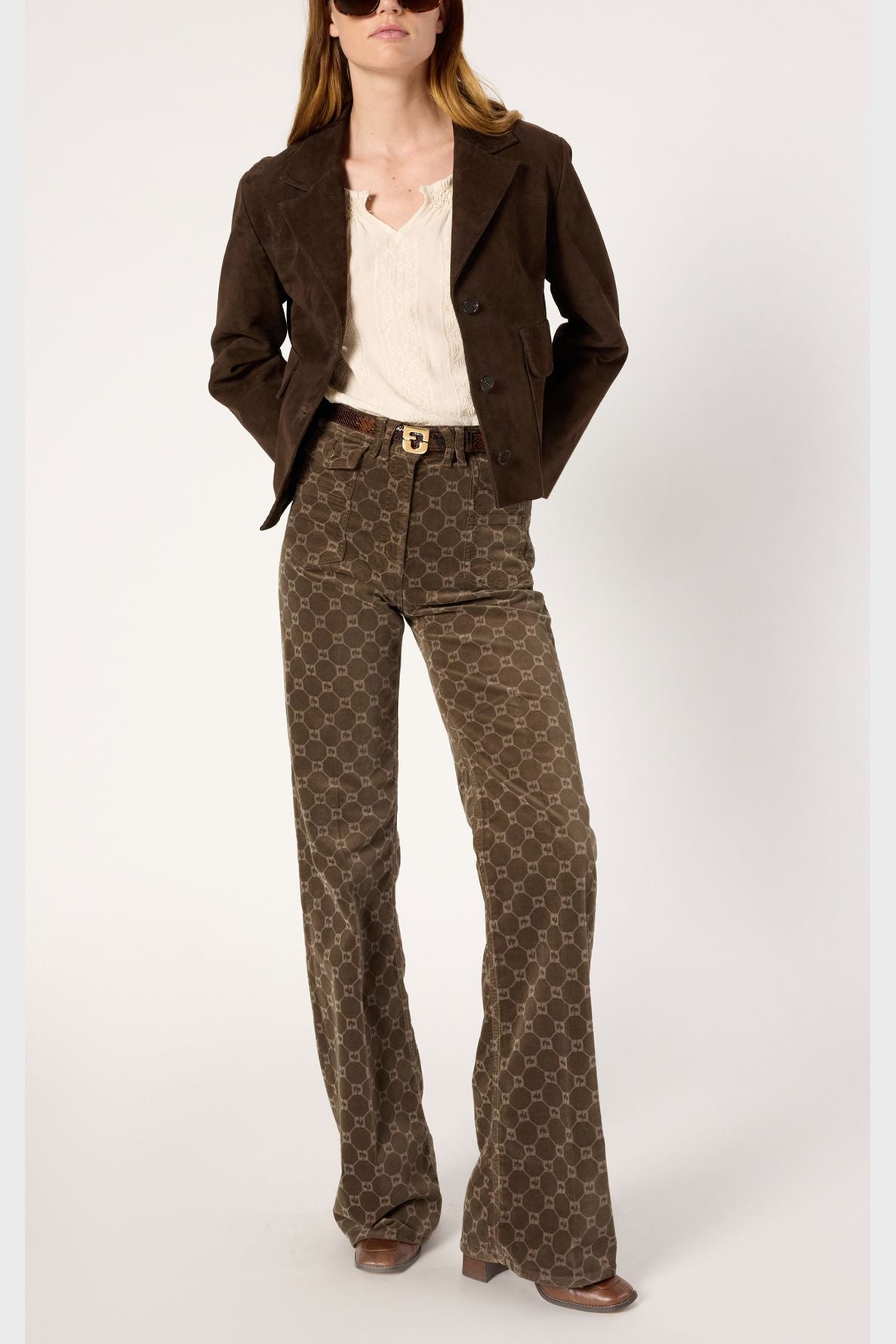 A woman wearing a brown suede jacket, cream blouse, brown patterned flare pants, brown belt with buckle, and brown shoes stands with hands in her pockets against a plain light background.