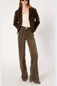 A woman wearing a brown suede jacket, cream blouse, brown patterned flare pants, brown belt with buckle, and brown shoes stands with hands in her pockets against a plain light background.