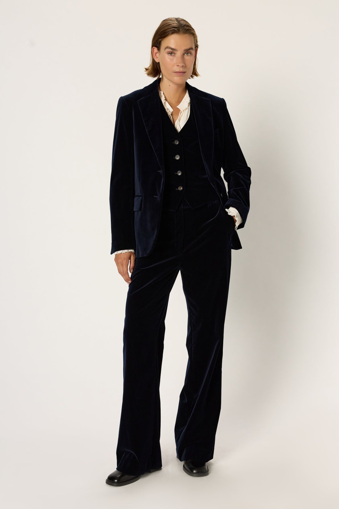 Person with short hair wearing a bleu nuit velvet suit featuring a pantalon en velours à coupe large, black vest, white shirt, and black shoes stands confidently against a plain light background.