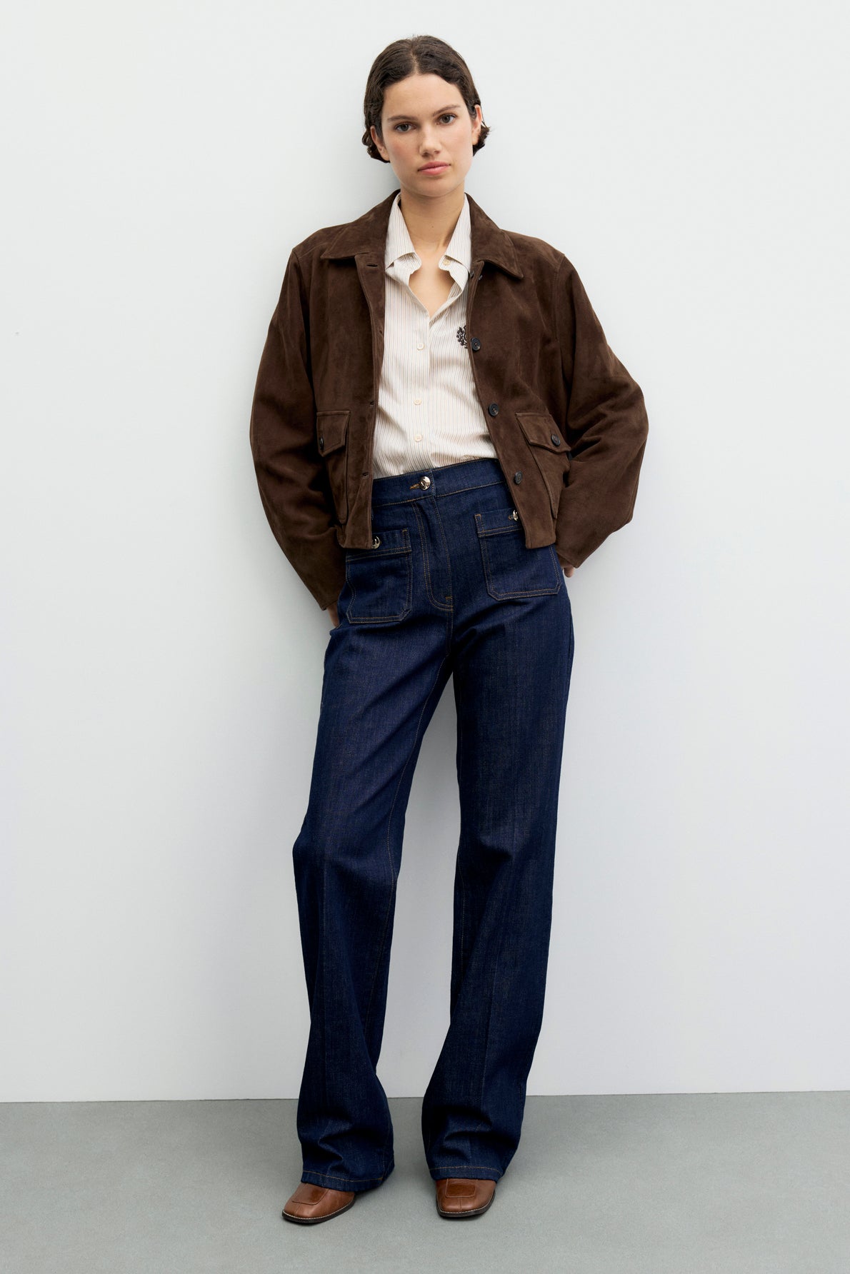 Person standing against a white wall, wearing a brown suede jacket over a light-colored shirt, high-waisted dark blue jean brut à coupe droite, and brown boots. Hands are in pockets and expression is neutral.