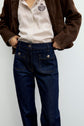 A person wearing a brown jacket, a white pinstripe shirt with a crest on the chest, and a jean brut taille haute à coupe droite with large front pockets stands against a light background.