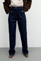 Person wearing dark blue wide-leg jeans with front patch pockets in a coupe droite silhouette, paired with a beige shirt and a brown jacket. Their hands are in their pockets, and they are standing on a gray floor against a plain white wall.