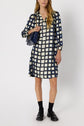 Woman wearing a knee-length, long-sleeve robe imprimé géométrique with a navy blue and white checkered pattern, black loafers, black socks, and holding a navy quilted bag, standing against a white background.