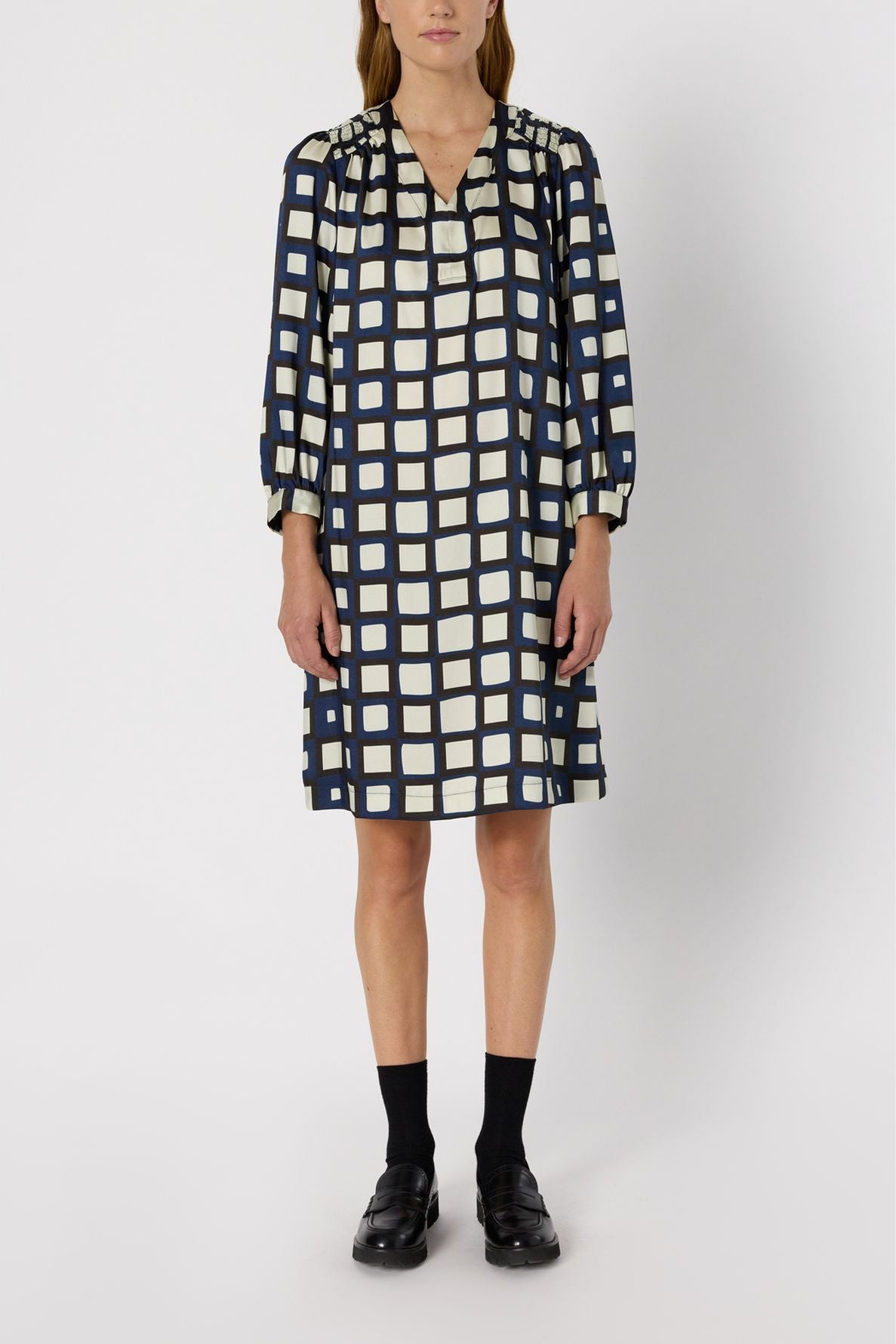 A person wearing a knee-length, long-sleeved robe courte femme with a navy blue and white checkered pattern reminiscent of a robe imprimé géométrique, paired with black socks and black shoes, standing against a plain white background.