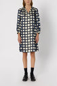 A person wearing a knee-length, long-sleeved robe courte femme with a navy blue and white checkered pattern reminiscent of a robe imprimé géométrique, paired with black socks and black shoes, standing against a plain white background.