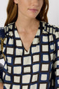 A woman wearing a patterned blouse with blue, black, and cream squares, featuring smocked shoulder details and a V-neck. Reminiscent of a robe imprimé géométrique, the look strikes a perfect balance between elegance and modern flair. She has light skin and brown hair, and carries a dark bag with a gold chain strap.
