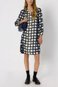A person wearing a navy and cream checkered robe courte femme with long sleeves, black loafers, black socks, and carrying a dark blue handbag, standing against a plain white background.