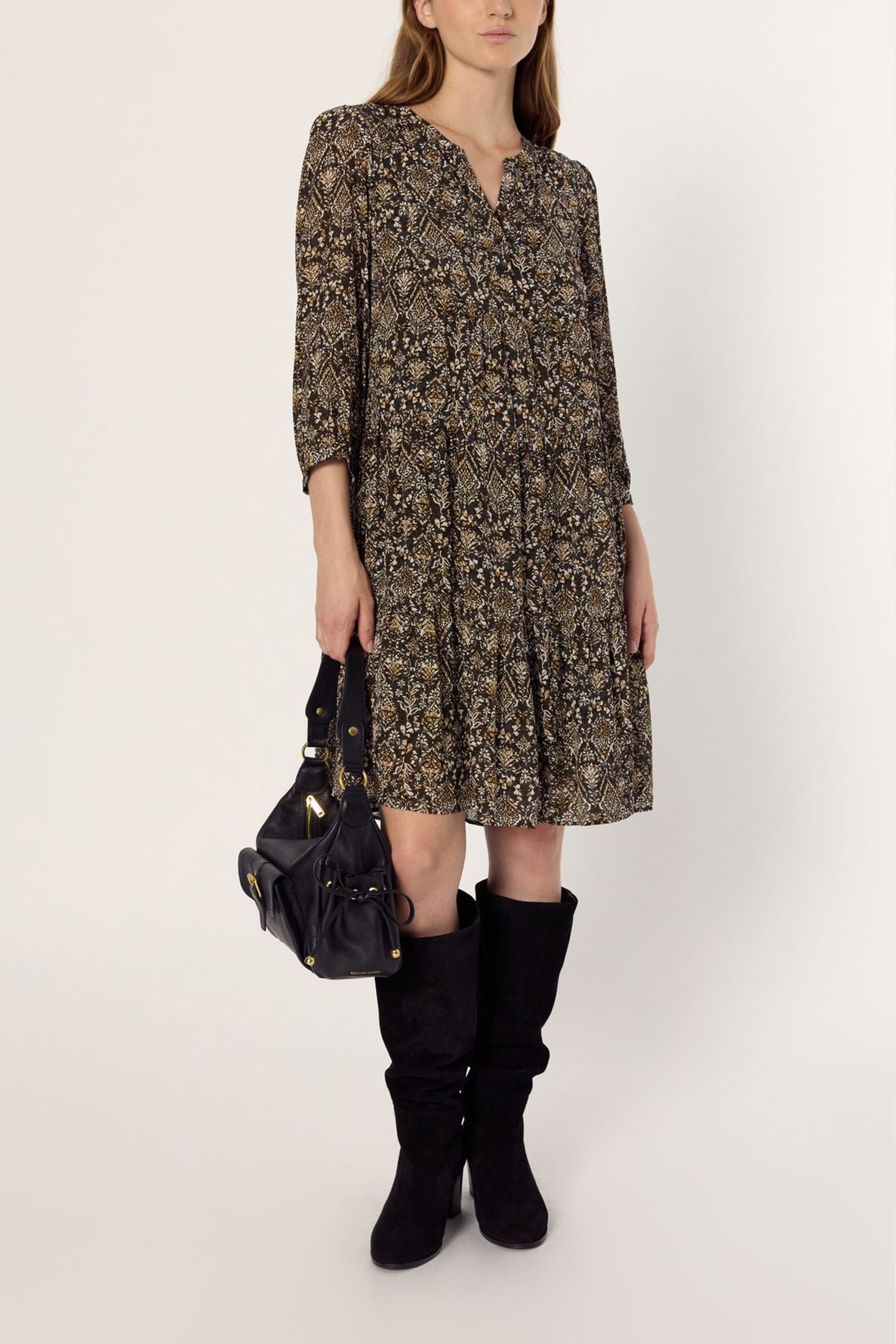 A woman in a robe bohème with motif ethnique, featuring long sleeves, holds a black handbag and wears black knee-high boots. She stands against a plain white background.