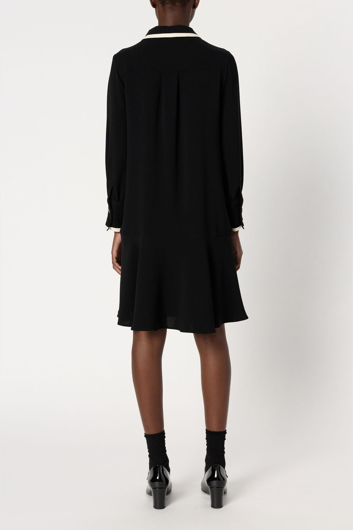 A person wearing a black long-sleeve robe courte chic with a flared hem, black socks, and black heeled shoes is standing facing away from the camera against a plain white background.