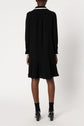 A person wearing a black long-sleeve robe courte chic with a flared hem, black socks, and black heeled shoes is standing facing away from the camera against a plain white background.