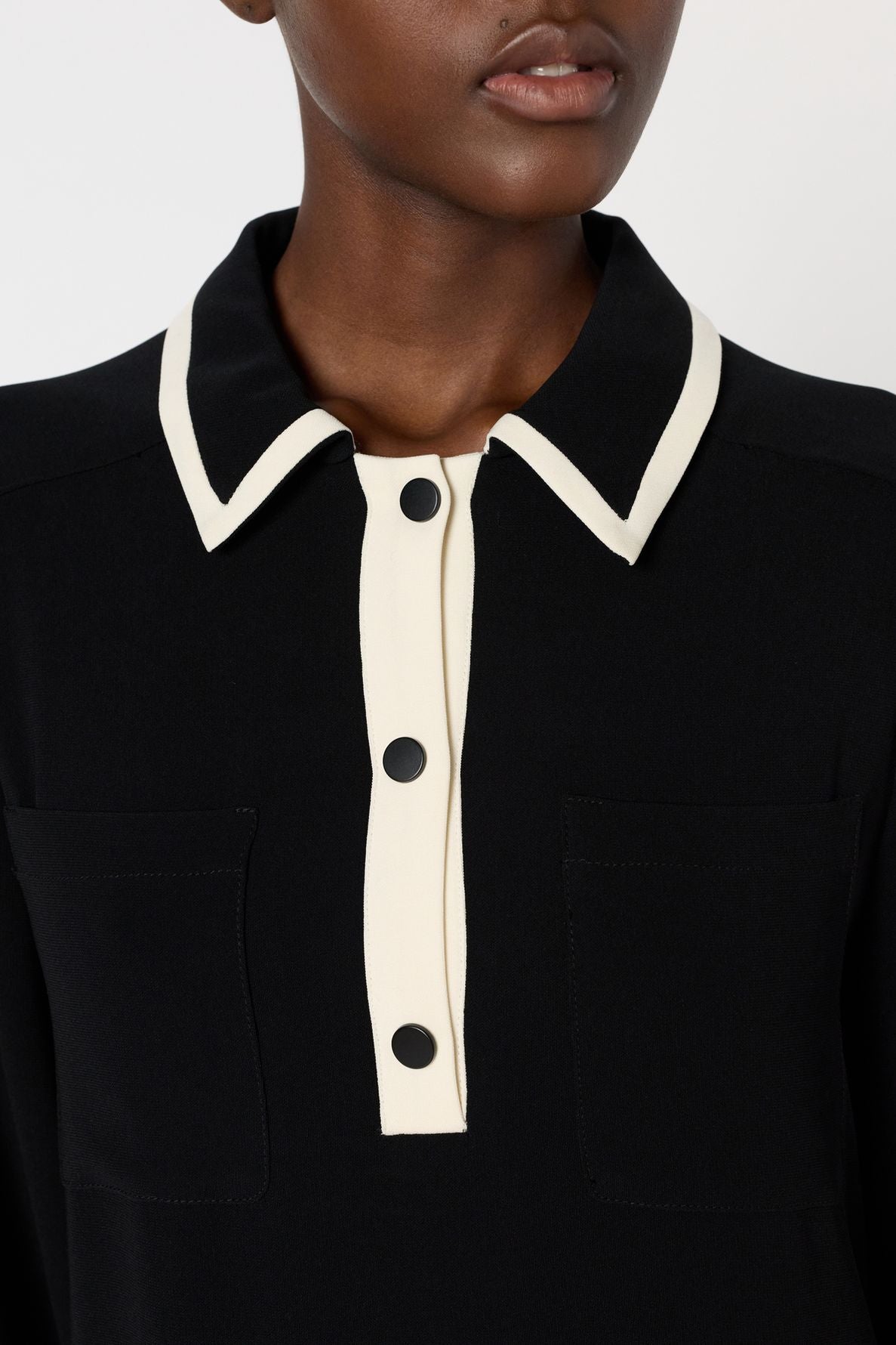 A person wearing a black collared shirt with white trim and black buttons, reminiscent of the elegant style of a robe col chemise. The shirt features two front pockets, and the image is cropped to show only the lower face and upper torso.
