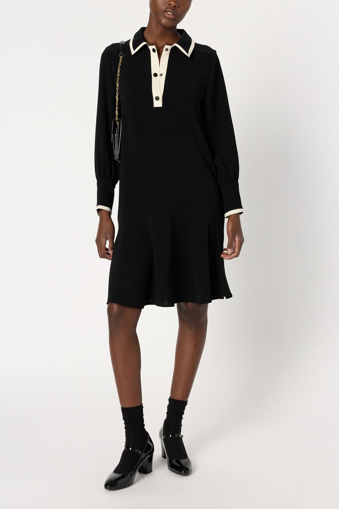 A person wearing a black robe courte chic, knee-length dress with a white col chemise collar and cuffs, black socks, black shoes, and carrying a black shoulder bag with a gold chain.