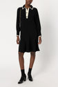 A person wearing a black robe courte chic, knee-length dress with a white col chemise collar and cuffs, black socks, black shoes, and carrying a black shoulder bag with a gold chain.