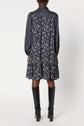 A person stands facing away, wearing a navy blue, long-sleeve, floral dress with a loose fit and ruffled hem, reminiscent of a robe mi-longue à col chemise. The look is completed with tall black boots.