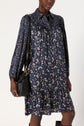 A person wearing a navy blue robe mi-longue à col chemise with a ruffled hem and floral print, holding a black shoulder bag, stands against a plain light background.