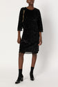 Person wearing a knee-length, black robe à sequins with three-quarter sleeves, black socks, and black heeled shoes, holding a bag with a gold chain strap, standing against a plain white background.