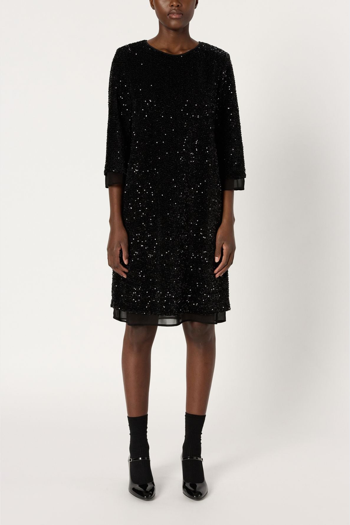 A person stands against a plain background wearing a black, knee-length robe à sequins with long sleeves, complemented by black socks and shiny black shoes. Their face is not visible.