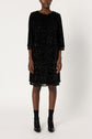 A person stands against a plain background wearing a black, knee-length robe à sequins with long sleeves, complemented by black socks and shiny black shoes. Their face is not visible.