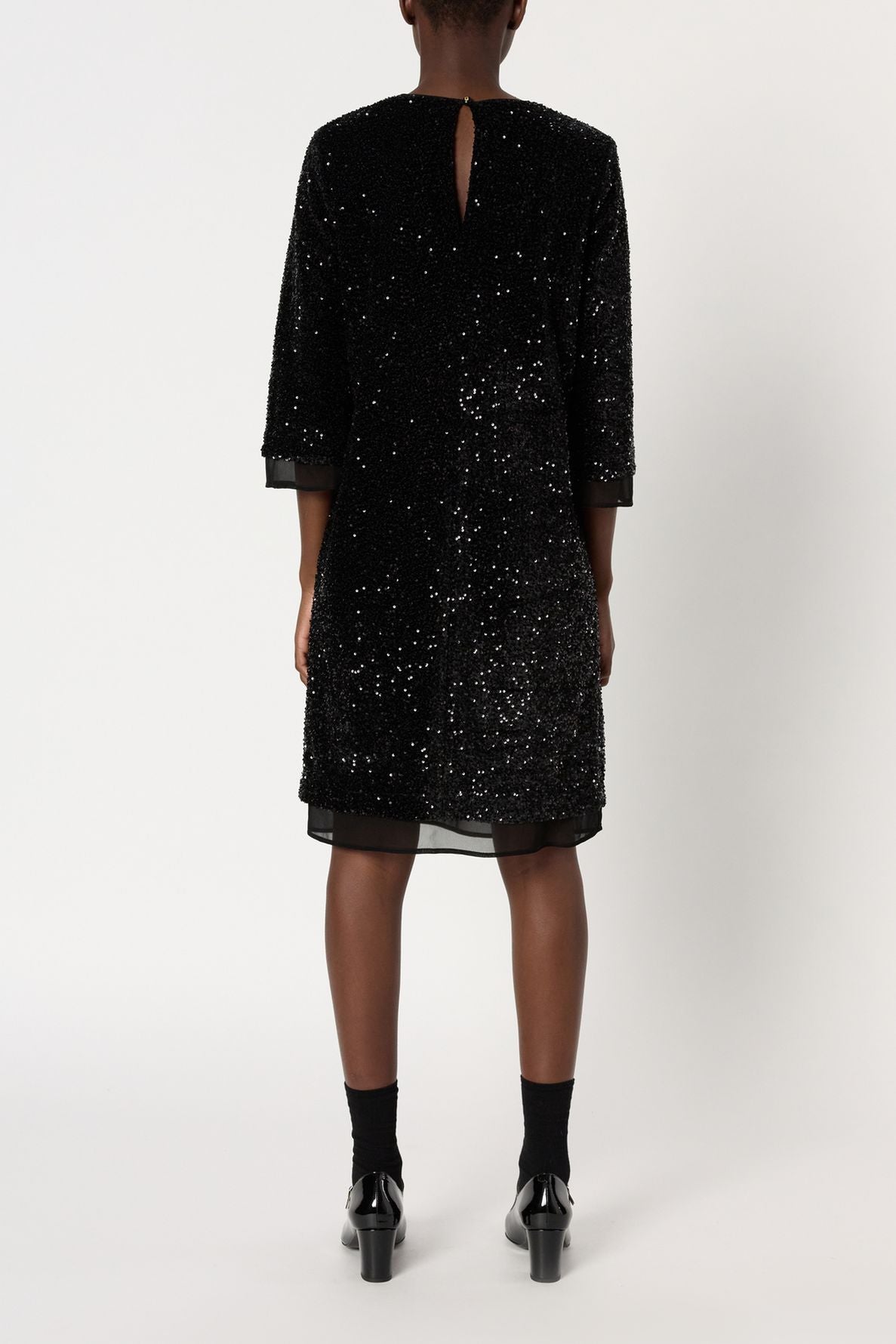A person stands facing away, wearing a robe à sequins noire mi-longue with three-quarter sleeves, black socks, and black shoes. The dress features a small keyhole opening at the top back. The background is plain white.