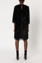 A person stands facing away, wearing a robe à sequins noire mi-longue with three-quarter sleeves, black socks, and black shoes. The dress features a small keyhole opening at the top back. The background is plain white.