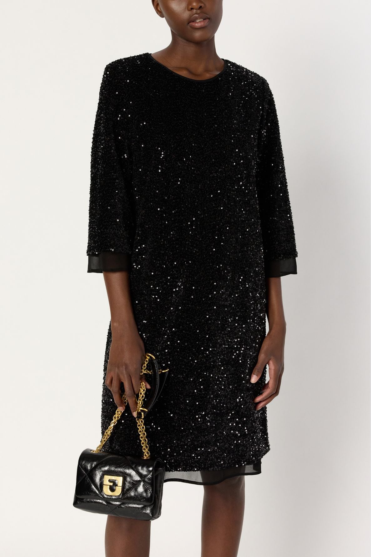 A person wearing a robe soirée noire à sequins, robe mi-longue aux manches trois-quarts, tient un petit sac à main noir matelassé avec une bandoulière chaîne et une boucle dorée. Le fond est entièrement blanc.