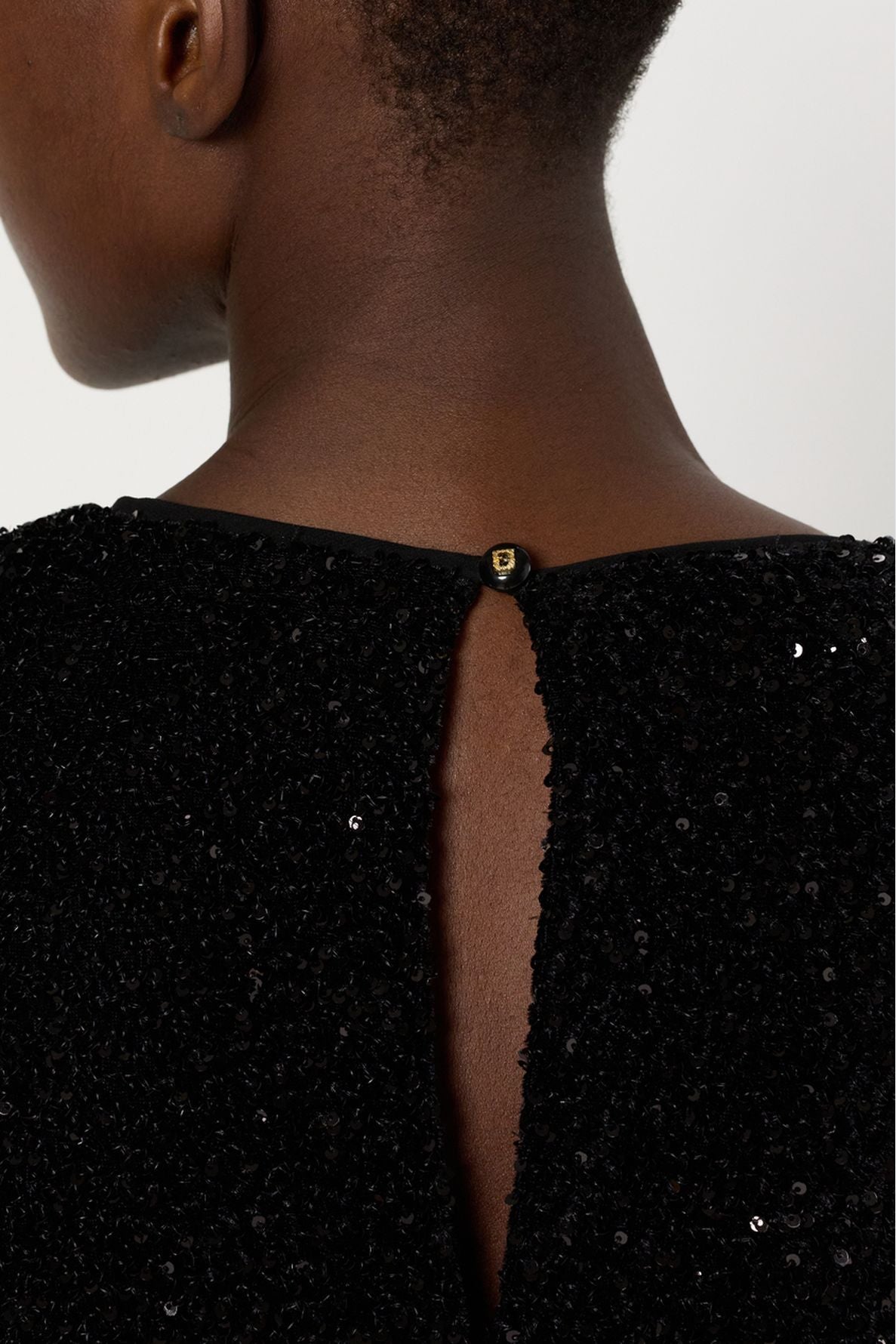 Close-up of the back of a person’s neck and shoulders, wearing a black robe à sequins with a small button fastening at the top of a back slit.