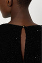 Close-up of the back of a person’s neck and shoulders, wearing a black robe à sequins with a small button fastening at the top of a back slit.