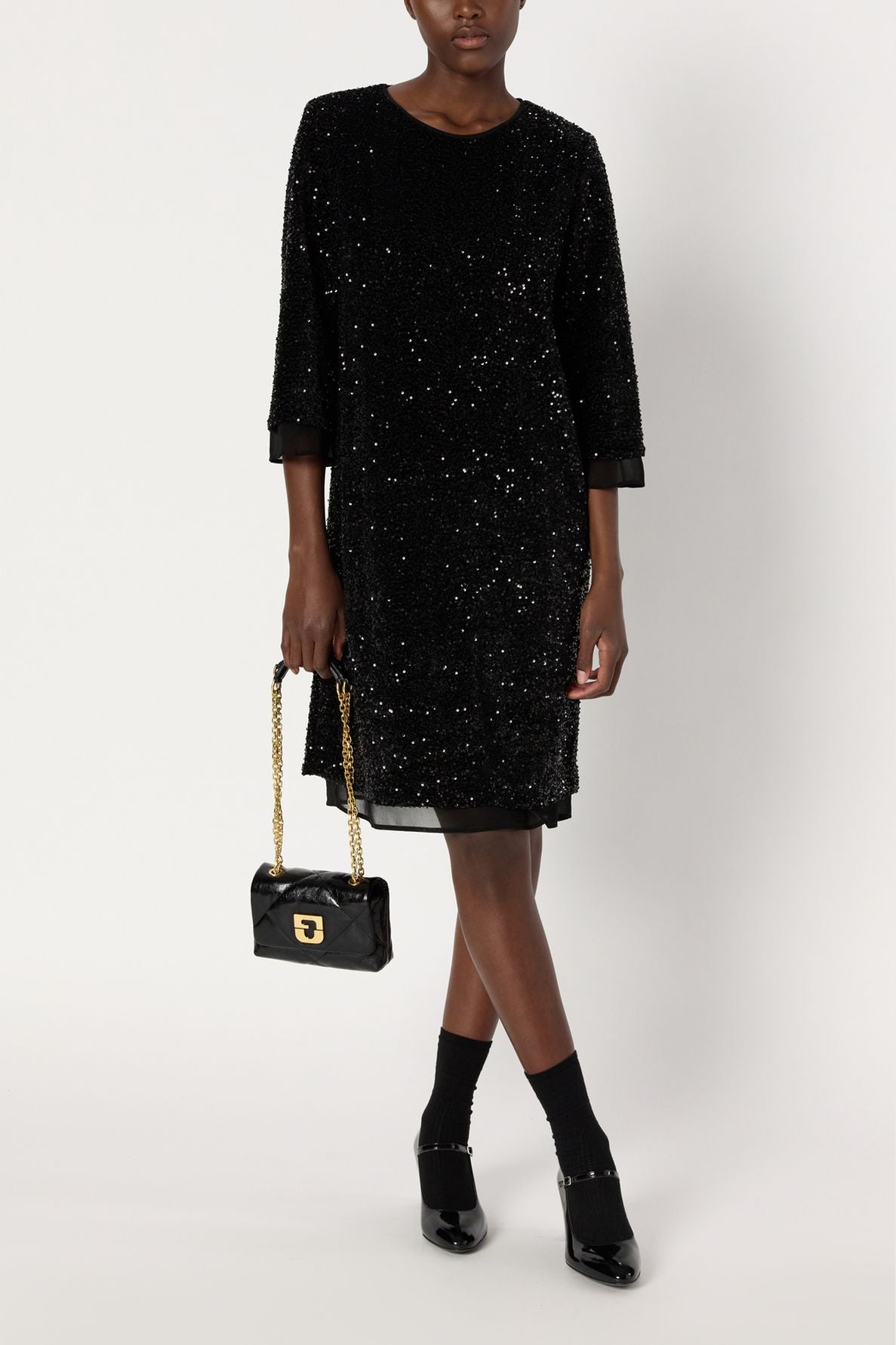 A person wearing a black sequined knee-length dress, or robe mi-longue à sequins, with black socks and black heeled shoes holds a small black handbag with a gold chain strap. The background is plain and white.