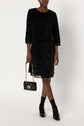 A person wearing a black sequined knee-length dress, or robe mi-longue à sequins, with black socks and black heeled shoes holds a small black handbag with a gold chain strap. The background is plain and white.