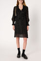 A woman wearing a robe mousseline noire, long-sleeved and knee-length with playful white pois lurex doré, styled with black loafers, black socks, sunglasses, and a chic black shoulder bag featuring a gold chain strap.