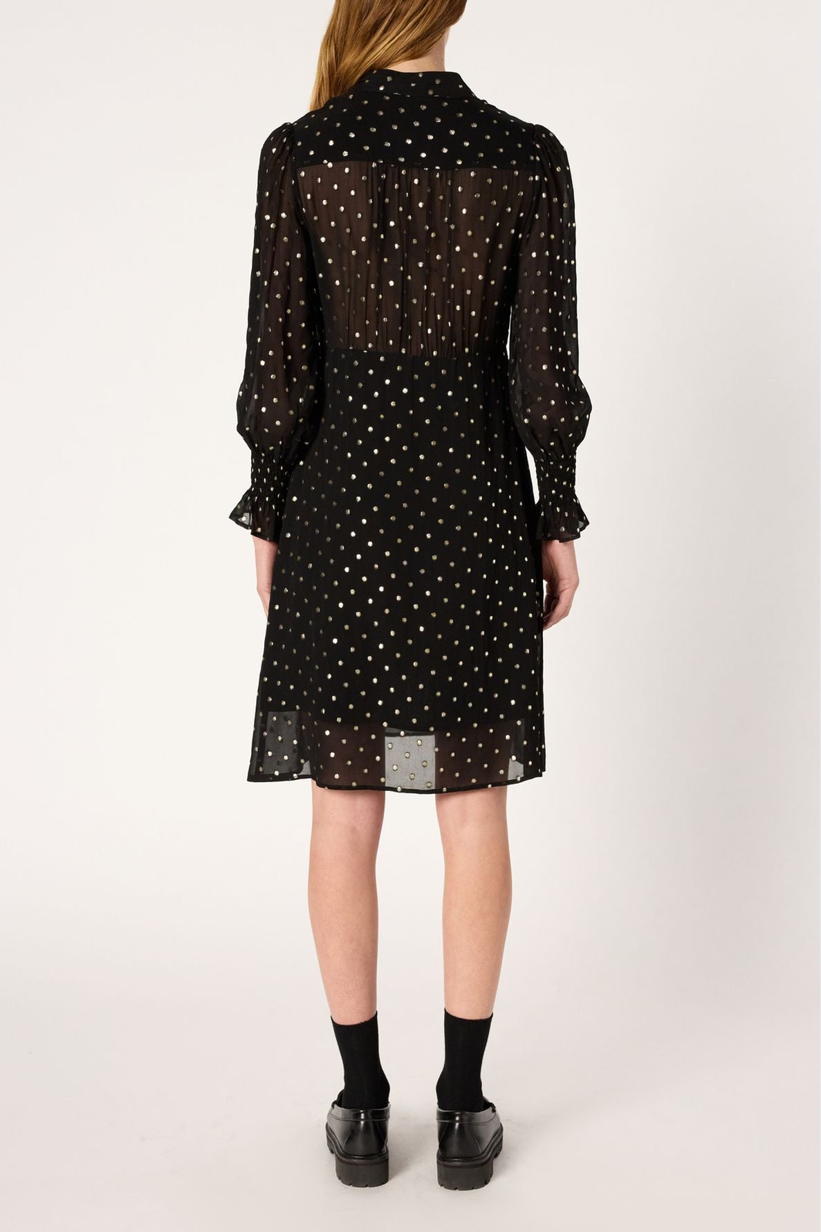 Person standing with their back to the camera, wearing a black, long-sleeve, knee-length robe mousseline noire with white pois lurex doré and a delicate col lavallière. The slightly sheer fabric subtly reveals a black underslip beneath, while the look is completed by black socks and black shoes.