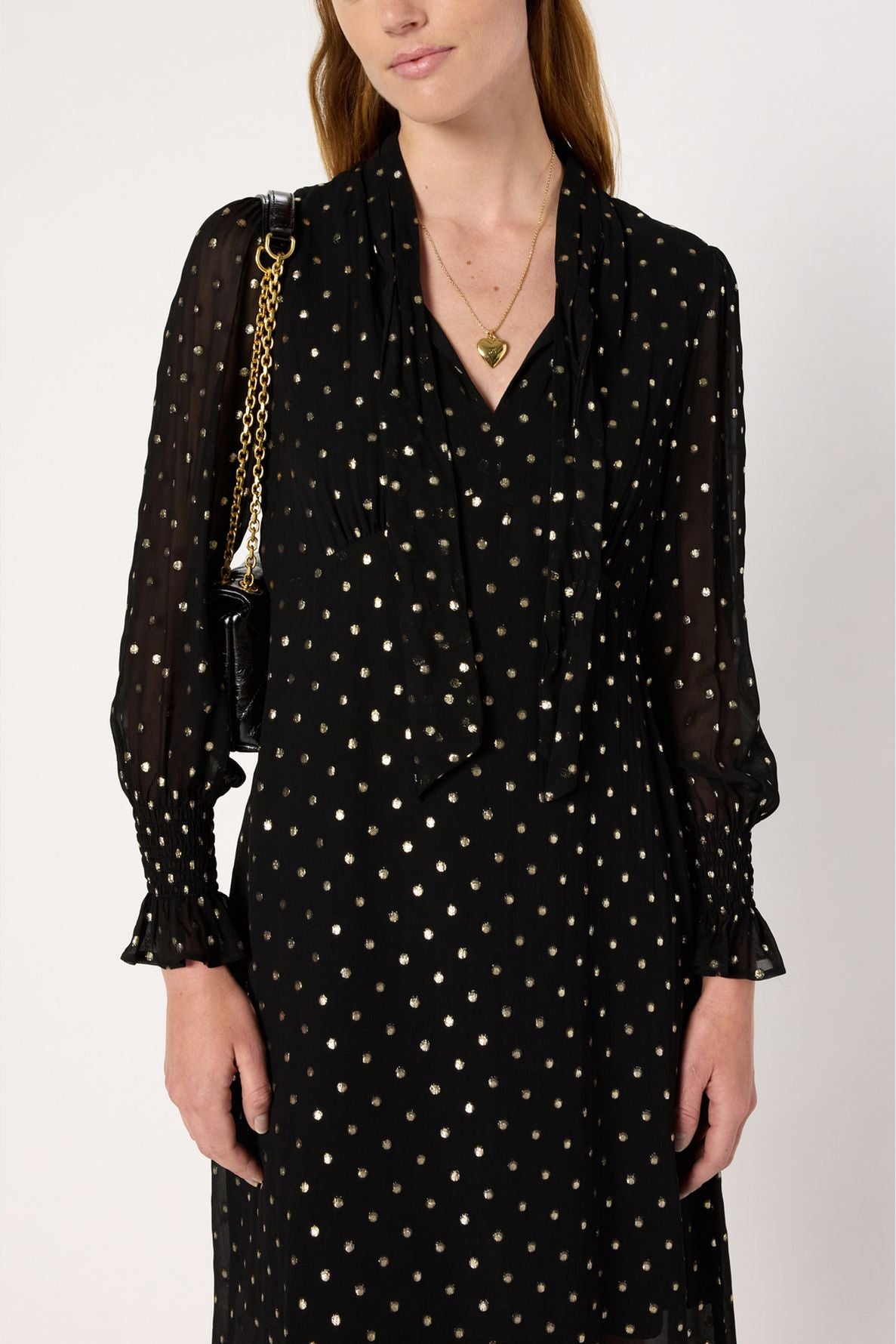 Woman wearing a robe mousseline noire adorned with pois lurex doré, featuring sheer long sleeves, a chic col lavallière neckline, a gold heart necklace, and a black shoulder bag with a gold chain strap.