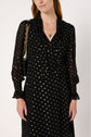 Woman wearing a robe mousseline noire adorned with pois lurex doré, featuring sheer long sleeves, a chic col lavallière neckline, a gold heart necklace, and a black shoulder bag with a gold chain strap.